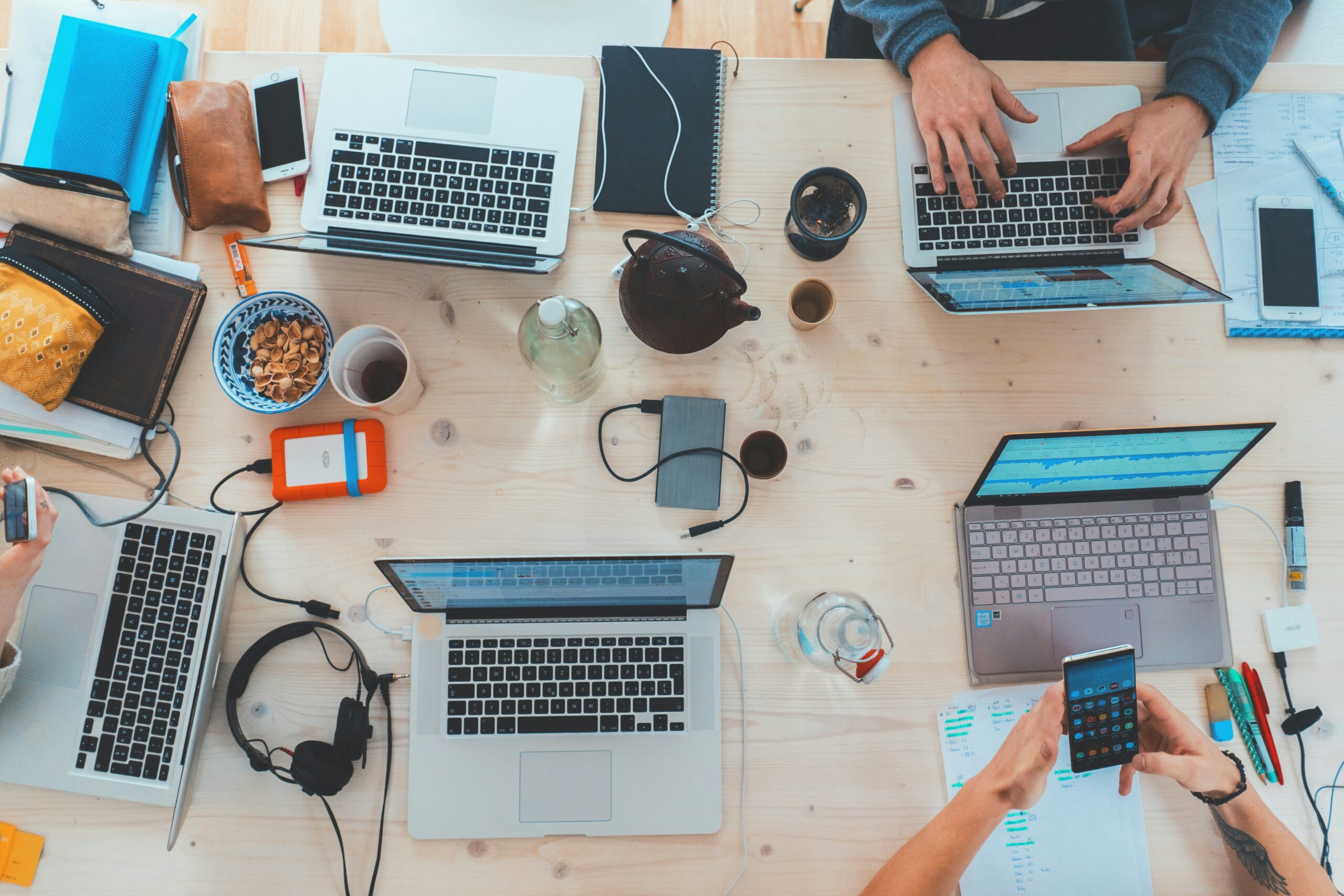 Top-down view of multiple laptops, phones, notebooks and work tools on a shared desk during a collaborative digital workspace session.