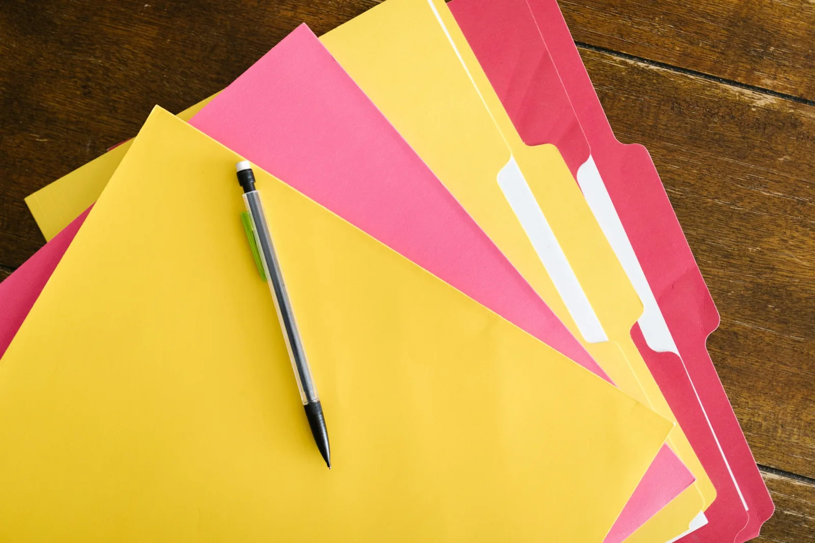 Yellow and pink folders arranged with a pen on a wooden desk.
